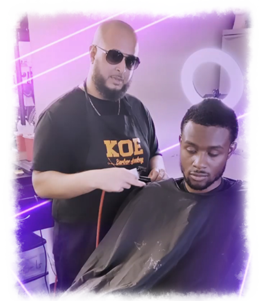 A barber in Bartow, wearing sunglasses and an apron emblazoned with "KOE," uses clippers to expertly cut the hair of a seated client. A ring light glows softly in the background.