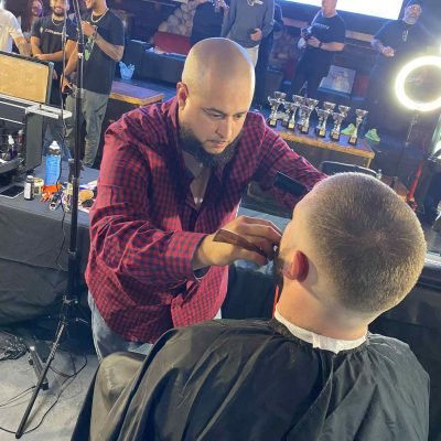 A barber in a red plaid shirt trims the beard of a seated client in Bartow, Florida. Several other people and trophies are visible in the background.