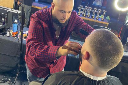 A barber in a red plaid shirt trims the beard of a seated client in Bartow, Florida. Several other people and trophies are visible in the background.