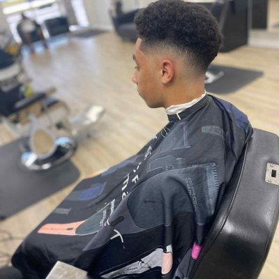 A person with short curly hair is sitting in a barber's chair inside a modern barbershop in Bartow, wearing a black barber cape.