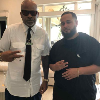 Two men stand indoors, facing the camera. The man on the left wears sunglasses, a white shirt, tie, and necklace. The man on the right wears a black shirt and makes a hand gesture. Both are posing casually, reminiscent of two cool customers at a barber in Bartow.