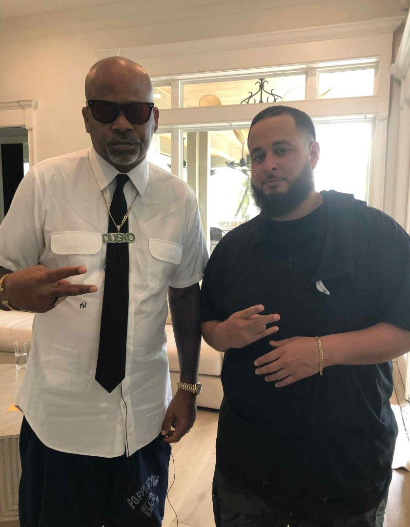 Two men stand indoors, facing the camera. The man on the left wears sunglasses, a white shirt, tie, and necklace. The man on the right wears a black shirt and makes a hand gesture. Both are posing casually, reminiscent of two cool customers at a barber in Bartow.