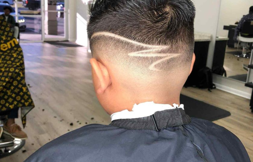 A person with short hair sits in a barbershop, showcasing a fresh undercut with artistic shaved lines on the back of the head. In the background, the shop's interior and another client can be seen. This stylish cut is expertly done by a barber in Bartow.