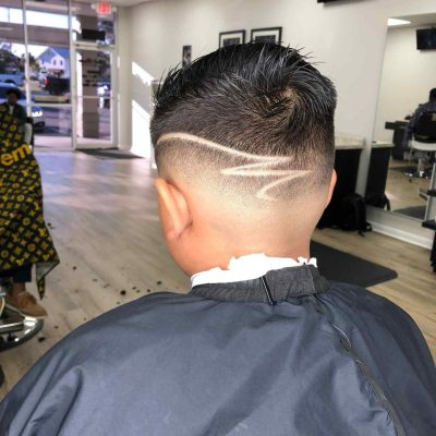 A person with short hair sits in a barbershop, showcasing a fresh undercut with artistic shaved lines on the back of the head. In the background, the shop's interior and another client can be seen. This stylish cut is expertly done by a barber in Bartow.