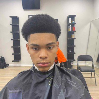 A young person with short, curly hair sits in a barber's chair, wearing a barber cape. The backdrop reveals shelves brimming with various hair products and a mirror, all under the skilled hands of a renowned barber in Bartow.
