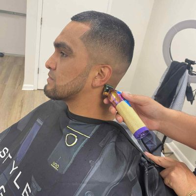 A man is getting a haircut at a barbershop in Bartow. The barber is using clippers to trim the hair around his ear. He is wearing a black barber cape, and a ring light is visible in the background.