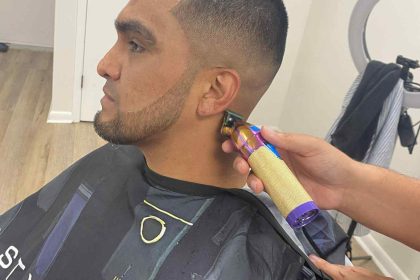 A man is getting a haircut at a barbershop in Bartow. The barber is using clippers to trim the hair around his ear. He is wearing a black barber cape, and a ring light is visible in the background.