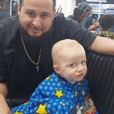 A child with a fresh haircut sits in a barbershop chair, covered with a colorful cape, while an adult stands beside, smiling. The busy Barber in Bartow shop buzzes with activity as other customers and barbers move in the background.