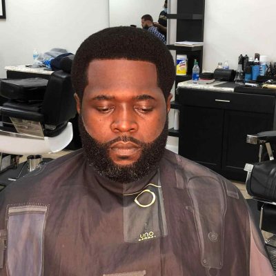 A man with a well-groomed beard and short haircut is seated in a Barber in Bartow chair, wearing a dark barber cape.