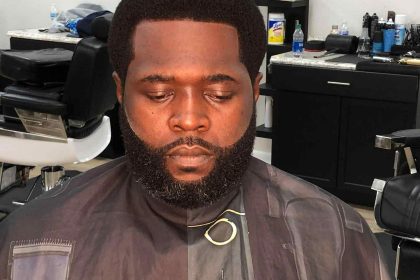 A man with a well-groomed beard and short haircut is seated in a Barber in Bartow chair, wearing a dark barber cape.
