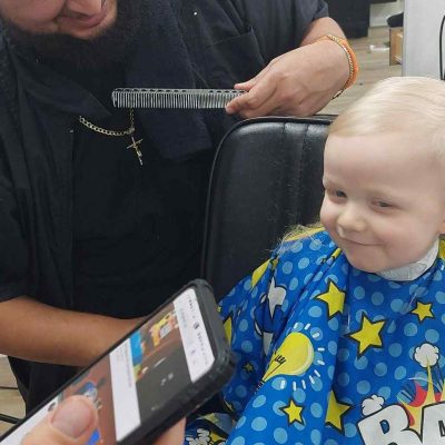 A Barber in Bartow cuts a young child's hair while the child, wearing a blue cape with cartoon characters, looks at a phone being held in front of them. The barber is smiling and holding a comb.