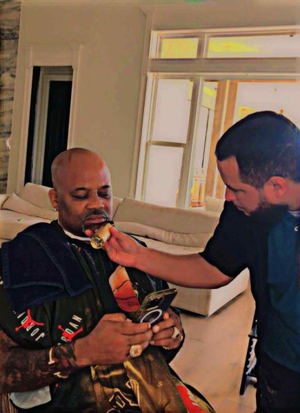 A bald man on his phone sits on a chair while another man in a black shirt assists him by holding a round object up to his chin. The scene reflects the precise care of a barber in Bartow, set against the backdrop of white walls and large windows.