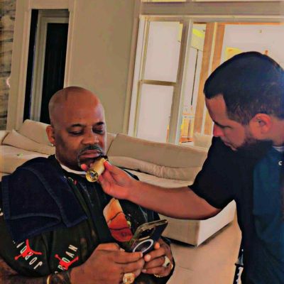 A bald man on his phone sits on a chair while another man in a black shirt assists him by holding a round object up to his chin. The scene reflects the precise care of a barber in Bartow, set against the backdrop of white walls and large windows.