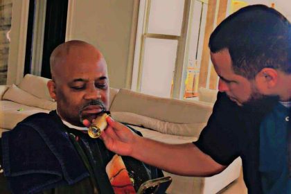 A bald man on his phone sits on a chair while another man in a black shirt assists him by holding a round object up to his chin. The scene reflects the precise care of a barber in Bartow, set against the backdrop of white walls and large windows.