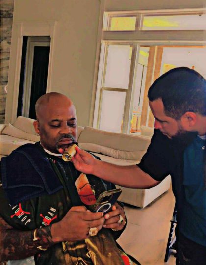 A bald man on his phone sits on a chair while another man in a black shirt assists him by holding a round object up to his chin. The scene reflects the precise care of a barber in Bartow, set against the backdrop of white walls and large windows.