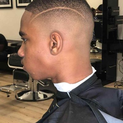 A person with a fresh fade haircut featuring a clean shaving line on the side, seated in a barbershop chair wearing a black cape. The Barber in Bartow works meticulously as mirrors and wall art adorn the background.
