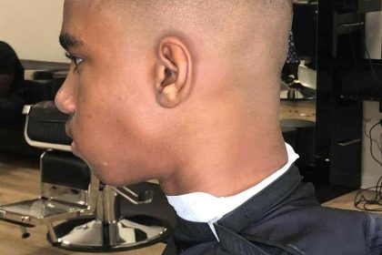 A person with a fresh fade haircut featuring a clean shaving line on the side, seated in a barbershop chair wearing a black cape. The Barber in Bartow works meticulously as mirrors and wall art adorn the background.