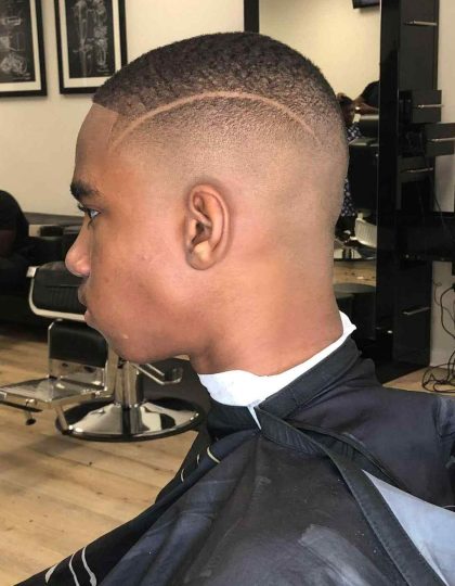 A person with a fresh fade haircut featuring a clean shaving line on the side, seated in a barbershop chair wearing a black cape. The Barber in Bartow works meticulously as mirrors and wall art adorn the background.