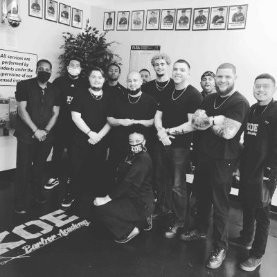 Group of twelve people in black attire posing together indoors, with framed photos on the wall and a plant in the background. The setting appears to be a barber academy in Bartow, Florida.