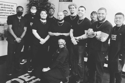 Group of twelve people in black attire posing together indoors, with framed photos on the wall and a plant in the background. The setting appears to be a barber academy in Bartow, Florida.